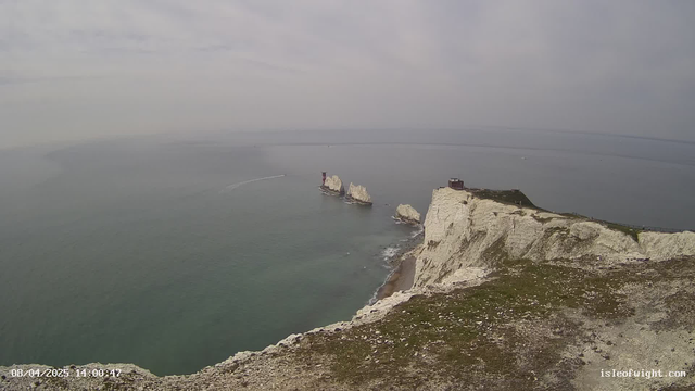 A view of a coastal landscape featuring white chalk cliffs on the right side. Below the cliffs, there is a rugged shoreline with some rocky outcrops. In the distance, several small rocky islands are visible on the water. A thin boat wake leads towards the islands, and the sea is calm with a gentle ripple. The sky is overcast with soft, muted colors, creating a hazy atmosphere. The timestamp at the bottom indicates the date and time of the image.