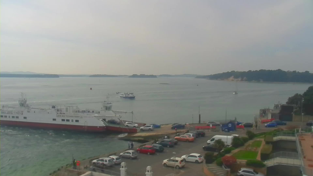 A view of a harbor with several boats on the water. In the foreground, a large white and red ferry is docked, labeled "GIVE WAY TO CHAIN FERRY." Several smaller boats are visible further out on the water. The shoreline features a parking area with various cars and a few people near the water. In the background, a green island is visible, along with a distant tree line and a cloudy sky.