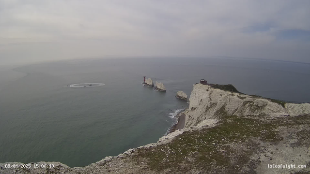 A coastal view featuring steep white cliffs along the shoreline. The sea is calm with gentle waves, reflecting a cloudy sky. In the distance, there are several rocky outcrops, including two lighthouses; one is painted red and white, while the other has a more natural appearance. Near the center, a group of small boats is creating circular patterns in the water. The foreground shows grass-covered land, suggesting a high vantage point overlooking the sea.