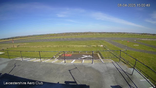 A clear blue sky with a few wispy clouds is visible above a grassy airstrip. In the foreground, there is a black and white helipad marked on the ground, featuring a white cross symbol. Adjacent to the helipad, there is a red marker or warning sign. The airstrip extends into the distance with neatly manicured grass on either side, and a wooden fence runs along the perimeter. The scene is bright and well-lit, indicative of a sunny day.