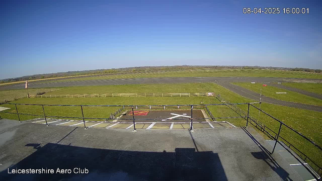 A clear blue sky stretches overhead, with bright sunlight illuminating a grassy airfield. In the foreground, a balcony railing is visible, with shadows cast on the ground. Below, the airfield features a green lawn surrounded by a wooden fence, and a marked helipad with a white “X” shape in the center. A small red sign can be seen near the edge of the helipad. In the distance, runways are visible, leading off into the horizon lined with trees. The scene conveys a sense of openness and tranquility at the Leicestershire Aero Club.