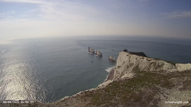 A panoramic view of the sea and coastal cliffs under a partly cloudy sky. The foreground features grassy terrain leading to steep white chalk cliffs. In the distance, several rocky formations, known as the Needles, rise from the water. Boats are visible on the water, leaving trails as they move. The ocean appears calm with shimmering reflections from the sunlight.