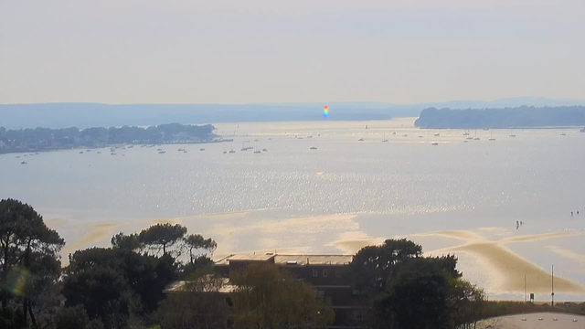A wide view of a tranquil coastal scene with calm waters reflecting sunlight. Small boats are scattered across the water, and a distant shoreline is visible, lined with trees. In the foreground, there are patches of sandy beach and groups of people walking along the shore. The sky is bright and mostly clear, with a slight haze on the horizon. A faint rainbow shimmer can be seen on the water surface.