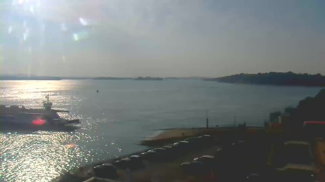 A serene view of a body of water under a bright sky. The sun is reflecting off the water creating sparkling highlights. In the foreground, there is a dock with a boat gently resting by the shore. There are several parked cars arranged neatly along the edge. In the distance, there are small islands visible and a gentle slope of land rises on one side of the water. The atmosphere is calm and peaceful.