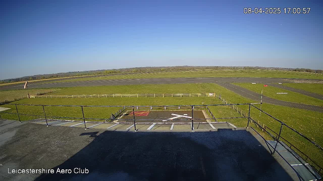 A panoramic view from a high vantage point overlooking a green airfield. In the foreground, there's a rooftop with a railing, partially shaded. Below, a grassy area features a white "X" marking on the ground, surrounded by a barricade. The airfield stretches out with runways visible and lined with a white fence. In the background, there are rolling green fields under a clear blue sky.