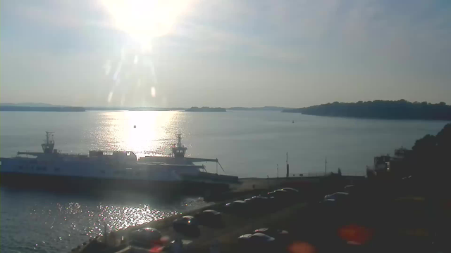 A bright sky with the sun shining directly above creates a reflective glow on the calm water of a bay. In the foreground, a white ferry is docked near a concrete pier lined with parked cars. The background features a distant shoreline with trees and hills, under a clear blue sky.