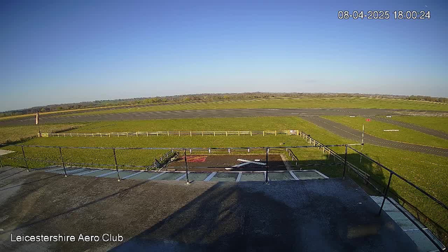 A clear blue sky stretches over a wide, grassy airfield. In the foreground, there is a concrete balcony railing with a view of the ground below. The airfield features a runway marked by white lines and a red windsock on a pole, indicating wind direction. A fenced area is visible along the edge of the field, surrounded by green grass. The scene is bright and devoid of clouds, suggesting a sunny day.