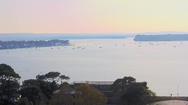 A serene view of a calm waterway with various small boats scattered across the surface. In the foreground, there are trees with dark green foliage, and a building with a flat roof partially visible. The background features a shoreline dotted with houses and more trees, extending into a soft gradient of colors in the sky, from pale pink to light blue. The distant land forms a gentle outline against the horizon. The overall scene conveys a tranquil and peaceful atmosphere.