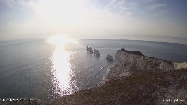 A serene coastal scene featuring calm waters reflecting sunlight. In the background, large rock formations rise from the sea, with one rocky outcrop supporting a small structure. A steep, white chalk cliff leads up to grassy areas, adding to the coastal landscape. The sky is bright with soft clouds, creating a peaceful atmosphere.