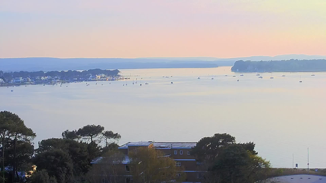 A serene landscape view of a calm body of water at sunrise. The water reflects soft pastel colors of pink and blue, with several small boats scattered across the surface. On the left, a line of houses and trees is visible along the shore. In the background, gentle hills rise against the horizon, creating a peaceful and tranquil scene.
