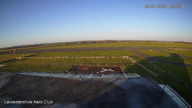 A clear blue sky stretches overhead, while a vast green field occupies the foreground. Visible in the foreground is a runway area with a white wooden fence outlining it. A red object, likely a vehicle, is parked along the edge of the runway. The ground shows signs of well-maintained grass, and the scene is illuminated by natural sunlight. In the lower left corner, there is a structure resembling an outdoor deck, with steps leading down from it. The timestamp in the top right corner reads 19:00 on April 8, 2025.