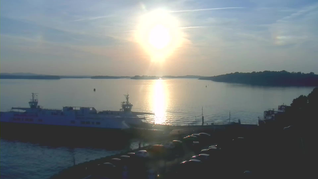 A serene view of a body of water at sunset. The sun is low in the sky, casting a bright reflection across the water's surface. A large ferry boat is docked on the left side, with cars parked nearby along the dock. In the background, there are wooded islands and hills silhouetted against the colorful sky. The atmosphere is calm and peaceful, with soft clouds scattered above.