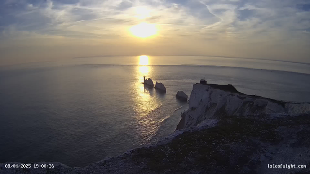 A scenic view of a coastline during sunset. The sun is low in the sky, casting a warm golden light over the calm sea. Silhouetted against the light are large white chalk cliffs and several rock formations protruding from the water. In the background, a faint horizon merges with the soft colors of the sky, which features wispy clouds. The scene conveys a tranquil atmosphere.