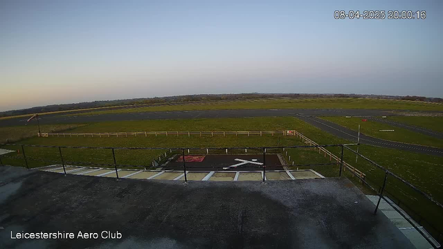 A panoramic view from a webcam at Leicestershire Aero Club. The image shows a wide expanse of green grassland, with a runway and taxiways visible in the foreground. In the distance, a fence outlines the area. A windsock is positioned on the left side, indicating wind direction. The sky above is clear to slightly overcast, reflecting the time nearing sunset. The date and time stamp in the corner indicates April 8, 2025, at 8:00 PM.