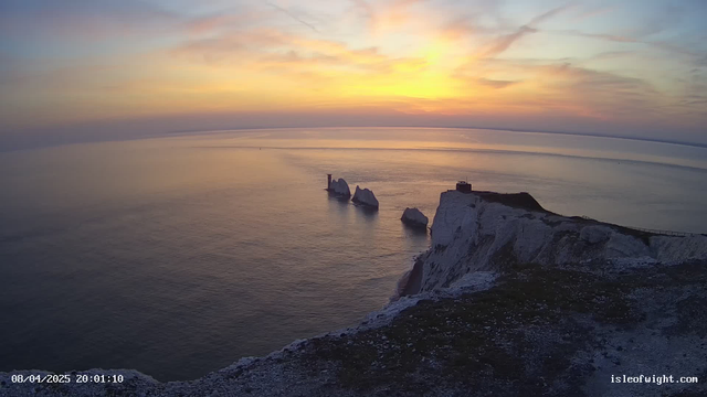 A wide view of the ocean at sunset, with soft pastel colors in the sky, including shades of orange, pink, and blue. In the foreground, there are white chalk cliffs leading down to the water. Three distinct rock formations jut out from the sea, with the largest on the left having a small building on top. The calm water reflects the colors of the sky, creating a serene atmosphere. The left side shows part of the cliff edge covered in grass and stones.