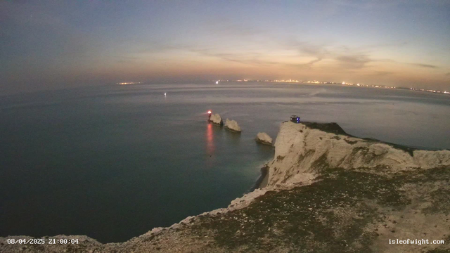 A panoramic view of a coastal landscape at dusk. In the foreground, rugged white cliffs extend into the water. The ocean is calm, reflecting soft hues of blue and purple from the sky. There are two white rock formations emerging from the water, with one featuring a small red lighthouse on top. In the background, faint lights from buildings or boats can be seen along the horizon. The date and time of the image are displayed in the bottom left corner.
