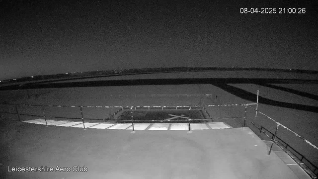 A dark, nighttime scene showing a view from a rooftop at the Leicestershire Aero Club. The foreground features a flat surface with a grid pattern created by light reflection. There are safety rails along the edge. In the background, faint lights from a distant landscape are visible, hinting at the presence of fields or buildings. The overall atmosphere is quiet and calm, with the time displayed indicating it is 21:00 on April 8, 2025.