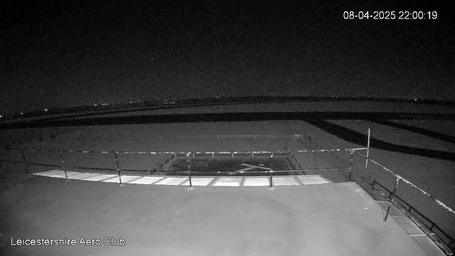 The image shows a dimly lit rooftop area with a railing and what appears to be a flat surface below, likely a hangar or a similar structure. The scene is predominantly dark, with a few lights visible in the distance suggesting the presence of other buildings or roads. The date and time in the corner indicate it is nighttime.