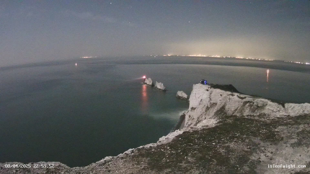 A night scene showing a coastline with white cliffs on the right side, leading to the ocean. In the ocean, there are jagged rock formations. A faint light comes from one of the rocks, and another light is visible further out in the water. The sky is dark, and stars are faintly visible. The horizon shows a distant glow from city lights, suggesting human activity. The foreground consists of a rocky area with some vegetation.