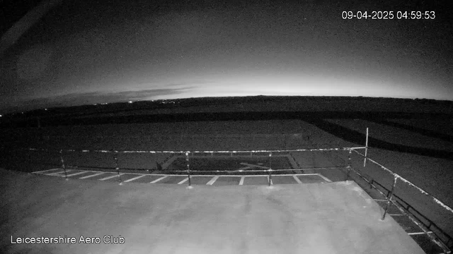 A dimly lit scene captured from a webcam at Leicestershire Aero Club, showing a flat, open area with an aircraft parking lot and a railing in the foreground. The sky is dark with subtle lightening on the horizon, indicating early dawn, while the landscape is mostly shadowed. The time displayed is 04:59:53 on April 9, 2025.