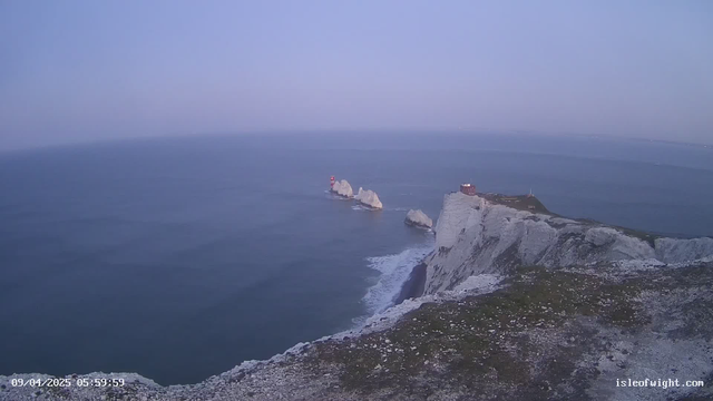 A coastal scene captured by a webcam showing cliffs with a grassy top. The ocean stretches out to the horizon, reflecting a soft blue hue. In the distance, there are two jagged rock formations with a red lighthouse on one of them. Waves gently break along the shoreline at the base of the cliffs. The sky is pale blue, indicating early morning light. A small building, possibly an observation point, is situated on the edge of the cliffs to the right. The date and time are displayed in the bottom left corner.