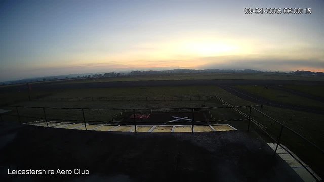 A panoramic view of an open field at dawn, with a soft gradient of colors in the sky transitioning from light blue to orange and yellow near the horizon. In the foreground, there is a rooftop with a railing overlooking a grassy area featuring a red surface and a white 'X' marking on it, indicating a possible landing spot. A gravel path is visible leading into the distance, and the horizon is lined with trees and hills, partially obscured by morning mist. The time stamp in the corner shows 06:00 on April 9, 2025.