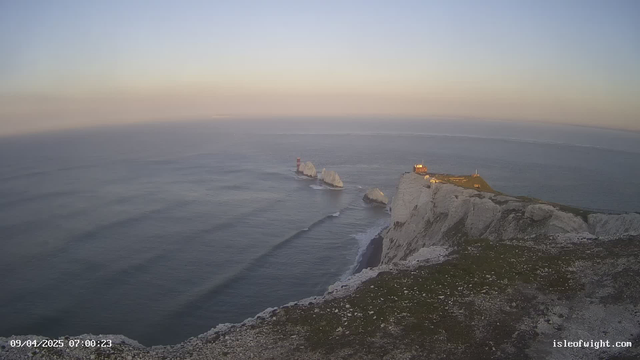 A coastal scene at dawn, featuring cliffs made of white chalk. In the water, several jagged rock formations with a lighthouse on one. The lighthouse is red and white, standing prominently against the soft morning light. The sky is a blend of pale blues and soft yellows as the sun rises. Gentle waves are visible in the calm sea. In the foreground, a grassy area leads to the cliff edge. The date and time are displayed at the bottom left of the image.