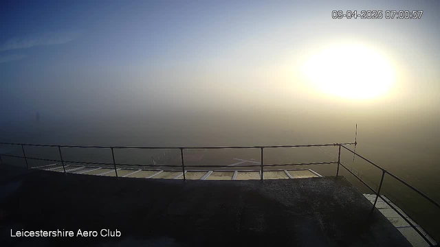 A foggy landscape viewed from a raised platform, with a blurred horizon and a bright sun rising in the distance. The foreground features a railing and a portion of a flat surface with grid-like patterns. Visibility is low due to thick fog, creating a soft, muted atmosphere. Time and date are displayed in the corner.