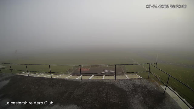 A foggy landscape from a webcam view at Leicestershire Aero Club. In the foreground, a railing surrounds a flat surface. Below, an airstrip with a small red area is barely visible through dense fog. The scene is muted with limited visibility, and the date and time, April 9, 2025, at 8:00:42, are displayed in the top right corner.