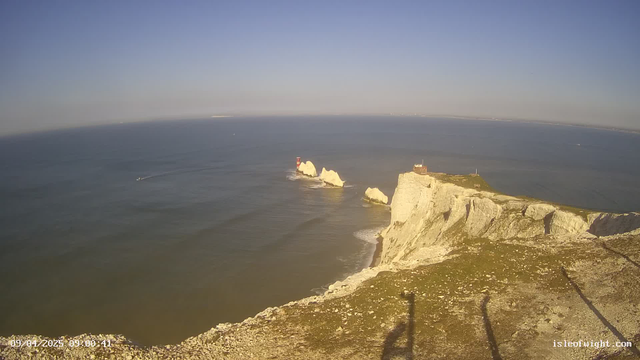 A scenic view of a coastal landscape featuring cliffs on the right side, with white rock formations jutting out into the water. In the sea, there is a small boat creating a wake as it moves toward the horizon. A red lighthouse stands on a rock in the middle of the water. The sky is clear and blue, with a few faint clouds, while the ocean appears calm with gentle ripples. The foreground shows grassy land leading up to the cliff's edge. The bottom of the image includes a timestamp indicating the date and time.