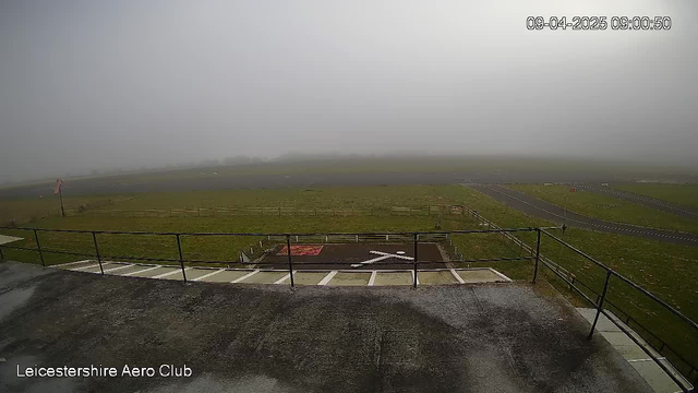 A foggy landscape from a webcam overlooking an open field. In the foreground, there is a railing and a marked area on the ground. The grass is damp and green, but visibility is limited due to the fog. There are outlines of structures and a road in the distance, but they are faintly seen through the haze. A sign with the name “Leicestershire Aero Club” is visible at the bottom. The date and time are displayed in the corner.