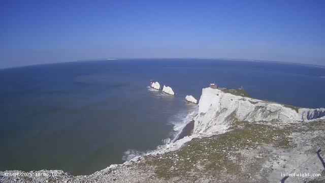 A panoramic view of the coastline featuring white chalk cliffs rising over a blue sea. In the distance, there are a few boats sailing on the water. The sky is clear with a few scattered clouds, and the horizon is visible where the sea meets the sky. Near the cliffs, two red and white striped lighthouses are positioned on the rocky shore, while a small building sits at the top of the cliffs. The foreground consists of grassy terrain and scattered stones leading towards the cliff's edge.