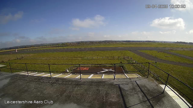 A view from a rooftop overlooking an airfield on a clear day. The sky is mostly blue with a few scattered clouds. In the foreground, there is a flat roof with a railing, and below, the airfield features a large green grass area bordered by wooden fences. A small area of the field is marked with a red square and a white cross. A paved runway is visible in the distance, lined with additional grass and a single road leading away from the airfield.