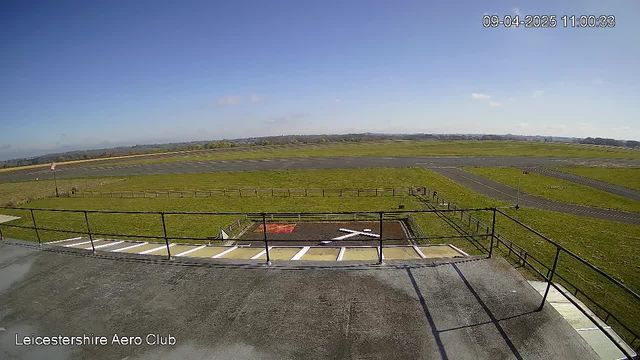 A clear sky stretches above a wide-open green field, interspersed with asphalt taxiways and a runway. In the foreground, there is a helipad marked with a large white cross and bordered by a fence. The scene is tranquil, with no visible aircraft or people, offering a peaceful view of the aerodrome.