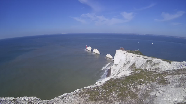 A panoramic view of a coastline featuring white chalk cliffs on the right, leading down to a blue sea. A small structure is visible atop the cliffs, while two rock formations with a red lighthouse stand in the water to the left. The sky is clear and blue with a few scattered clouds. Several small boats are seen on the water.