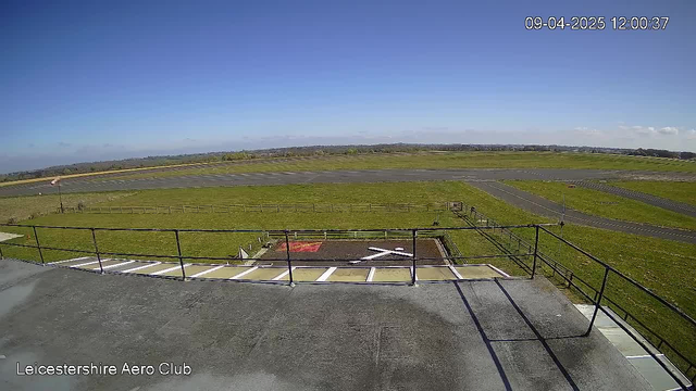 A clear sky with a few clouds overhead. The view shows a grassy area with a large open space, likely an airfield, featuring a runway in the distance. There are several wooden fences surrounding the grassy areas. In the foreground, there is a patio with a railing at the edge. A helicopter landing pad marked with a white cross is visible on the ground below, alongside some patches of red. The location is identified as Leicestershire Aero Club.