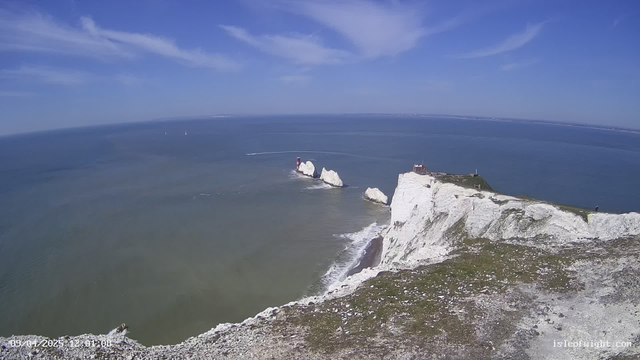 A coastal scene featuring cliffs with white chalk formations, overlooking a calm blue sea. In the distance, small sailboats are visible on the water. The sky is clear with a few scattered clouds. There is a red and white lighthouse situated on one of the cliffs, along with a building nearby. The foreground shows grassy terrain leading up to the cliff's edge.