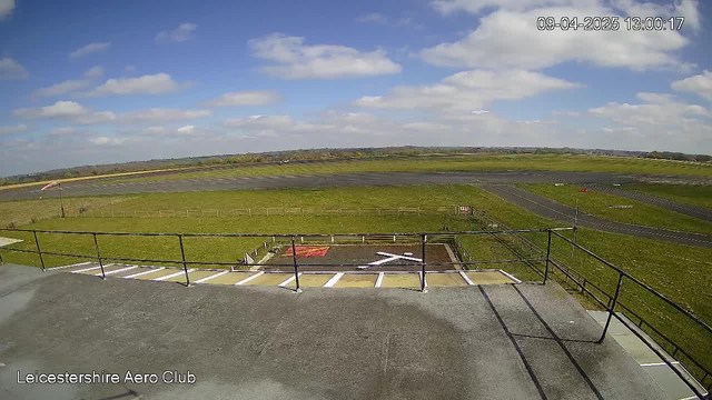 A panoramic view from a raised platform at the Leicestershire Aero Club. In the foreground, there is a concrete area with a white cross marking. Beyond it, a grassy expanse stretches out, bordered by a wooden fence. A runway can be seen in the distance, with a patch of tarmac on the left. The sky is mostly clear with a few clouds, indicating a bright day. The timestamp on the image shows the date and time as April 9, 2025, at 1:00 PM.