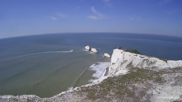 A panoramic view of a coastal landscape featuring white chalk cliffs on the right side, with a sandy beach at the base. The sea is a mix of green and blue hues, with gentle waves lapping at the shore. Two white rock formations protrude from the water, and a red and white lighthouse stands on one of the rocks. A small motorboat is visible, creating a spray as it moves across the water. The sky is bright blue with a few wispy clouds, and the horizon stretches far into the distance.