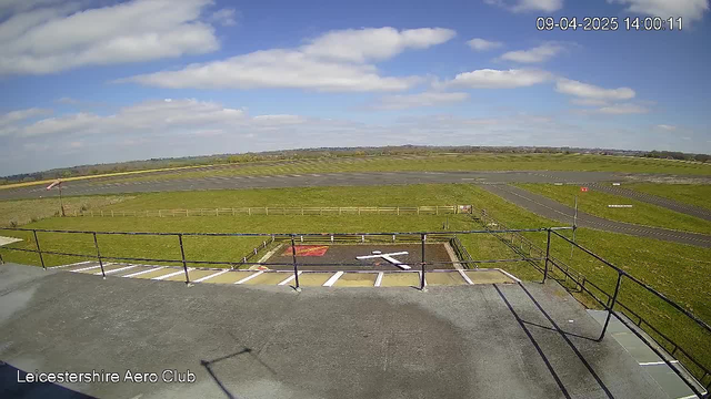A clear sky with scattered clouds is visible above a grassy area and an airfield. The foreground features a railing with a landing area marked by a white X on a black surface. In the distance, a runway can be seen, bordered by a wooden fence and patches of grass. The terrain extends towards low hills under a bright, sunny sky. The time and date are displayed in the top right corner of the image.