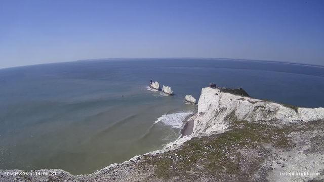 A panoramic view of a coastline featuring white chalk cliffs along the shore. The cliffs rise sharply from the water, with several rocky outcrops visible in the ocean. A small lighthouse is situated on one of the outcrops, painted in red. The sea is calm, displaying shades of blue and green, with gentle waves lapping at the shore. The sky is clear and blue, with a few wispy clouds, indicating a sunny day.