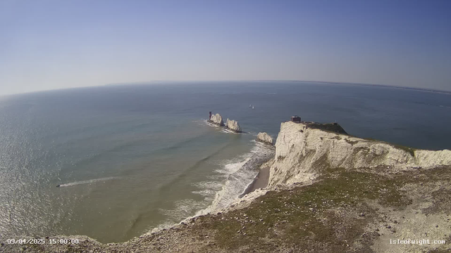 A scenic coastal view featuring white chalk cliffs along a shoreline. The sea is calm, with gentle waves lapping against the shore. In the distance, several small sailing boats are visible on the water. The sky above is clear and blue, indicating a sunny day. The land is grassy at the top of the cliff, while the beach below has a mix of sand and pebbles.