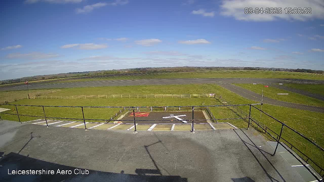 A view from a high vantage point at Leicestershire Aero Club. The scene shows a vast grassy area with a runway in the background. A helipad marked with a white cross is visible, along with a red area nearby. A wooden fence surrounds the field, and there are a few scattered clouds in a blue sky. The image is well-lit with sunlight, providing a clear view of the landscape and runway.