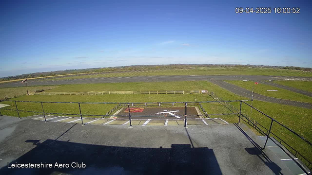 A view from a webcam high on a building at Leicestershire Aero Club, showcasing a clear blue sky with few clouds. In the foreground, a helipad is marked with a white 'H' symbol on a dark surface, surrounded by a flat grassy area. A wooden fence borders the grassy field, while a runway stretches into the distance, leading to more green fields and outlines of hills on the horizon. A windsock is visible on the left side of the image, indicating the wind direction. The scene is bright and open with no aircraft present. The time displayed is 16:00 on April 9, 2025.