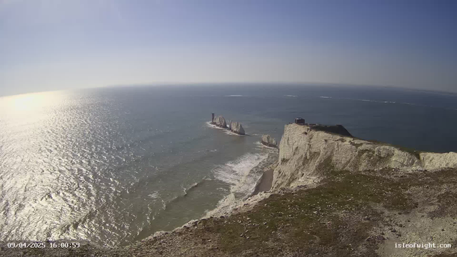 A panoramic view of the ocean and cliffs. The scene includes a bright blue sky with a few faint clouds overhead. Sunlight reflects off the water, creating a shimmering effect. To the right, a steep white cliff rises sharply from the water's edge, adorned with patches of green grass and rocks. In the background, several large rock formations emerge from the sea. A small structure can be seen atop the cliff, overlooking the water. The shoreline curves gently, with waves lapping at the base of the cliffs.