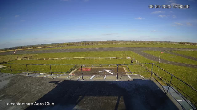 A wide view of an airfield on a clear day, with a blue sky and scattered clouds. In the foreground, there is a helipad marked with a white cross and surrounded by a fence. A grassy area surrounds the helipad and extends to the runway, which has a dark surface and is lined with white markings. In the distance, additional grassy fields and trees are visible. A single person is standing near the helipad, and a windsock is seen on the left side, indicating wind direction.