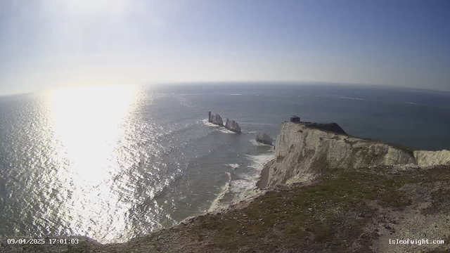 A coastal scene featuring a rocky shoreline with several large white chalk cliffs rising steeply from the water. In the middle distance, there are three distinctive rock formations emerging from the ocean. The sun is shining brightly, creating a shimmering reflection on the water's surface. The sky is mostly clear with a few scattered clouds. In the foreground, a grassy area with some rocky terrain is visible. The time stamp in the bottom left corner indicates the date and time of the image.