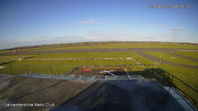 A panoramic view from a webcam at the Leicestershire Aero Club. The image features a vast expanse of green grass and a runway. In the foreground, there is a railing with a slight shadow. A windsock is visible to the left, indicating wind direction. In the distance, the sky is mostly clear with a few scattered clouds. A section of the runway is marked with white lines and there's a red and white sign visible on the right side. The time and date in the corner indicate it is 18:00 on April 9, 2025.