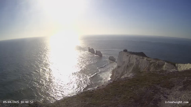 A bright sunny scene shows the ocean glistening under the sunlight. In the foreground, there are rocky cliffs with a grassy area and some vegetation. The sea shows gentle waves, and several large rock formations emerge from the water. In the distance, the horizon blends into a light blue sky. A structure is visible on top of the cliff to the right. The image includes a timestamp at the bottom left corner indicating the date and time.