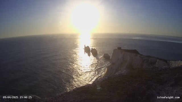 A panoramic view of the sea at sunset, with the sun's bright rays reflecting off the water, creating a shimmering path across the surface. In the foreground, jagged rocks emerge from the sea, surrounded by gentle waves. The sky is a gradient of soft blues and warm golds, fading into a clearer horizon. On the right, a light-colored cliff rises above the water, with a small structure on top. The bottom of the image displays the date and time.